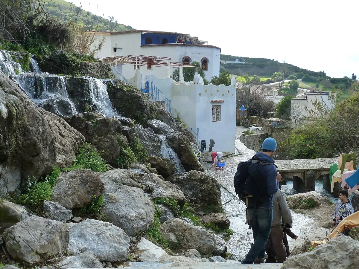 Le ruisseau de Ras El Maa en contrebas : l'eau dégringole de bloc en bloc sur de grosses pierres grises couvertes de mousse, la ruelle pavée en marches la longe, une habitante accroupie devant une bassine bleue au bord de l'eau, et deux marcheurs qui descendent l'escalier