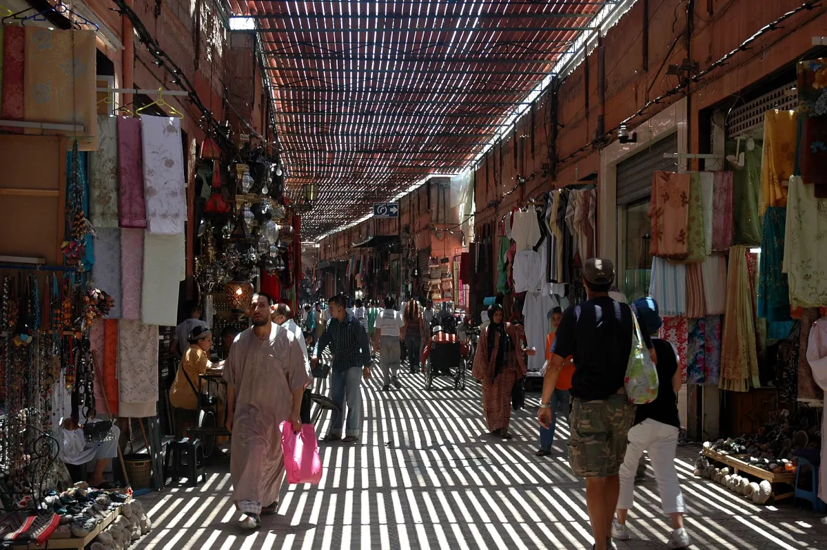 Allée couverte de la médina de Marrakech : bandes de lumière tombant à travers le treillis du toit, étoffes suspendues de part et d’autre et passants dans la pénombre rayée