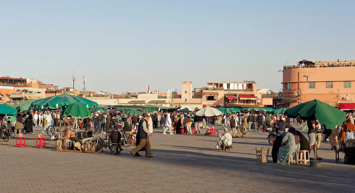 La place Jemaa el-Fna en fin d’après-midi : parasols verts des échoppes, foule dense, mobylettes et terrasses garnies sur les toits, entre les façades ocre