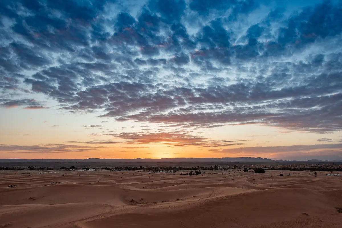 Depuis une crête de l'erg Chebbi, le soleil posé sur l'horizon d'une plaine sous un ciel très nuageux, les campements alignés au fond